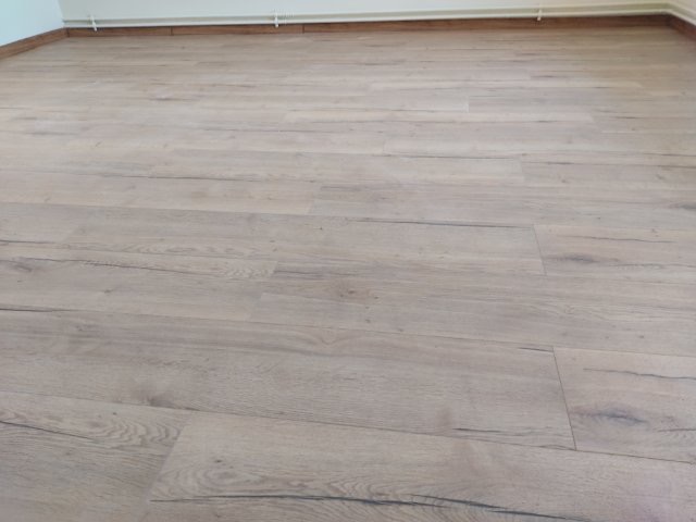 Jasne panele podłogowe imitujące drewno, ułożone w przestronnym pokoju z białymi ścianami i brązową listwą przypodłogową, widoczne łączenia paneli.