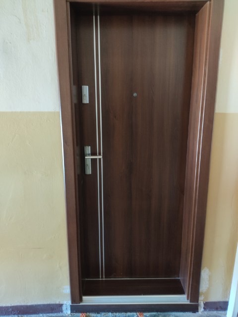 Ciemnobrązowe drzwi wejściowe z dwoma pionowymi, srebrnymi paskami, wizjerem i zamkiem, osadzone w brązowej futrynie w bloku mieszkalnym z żółto-białymi ścianami.