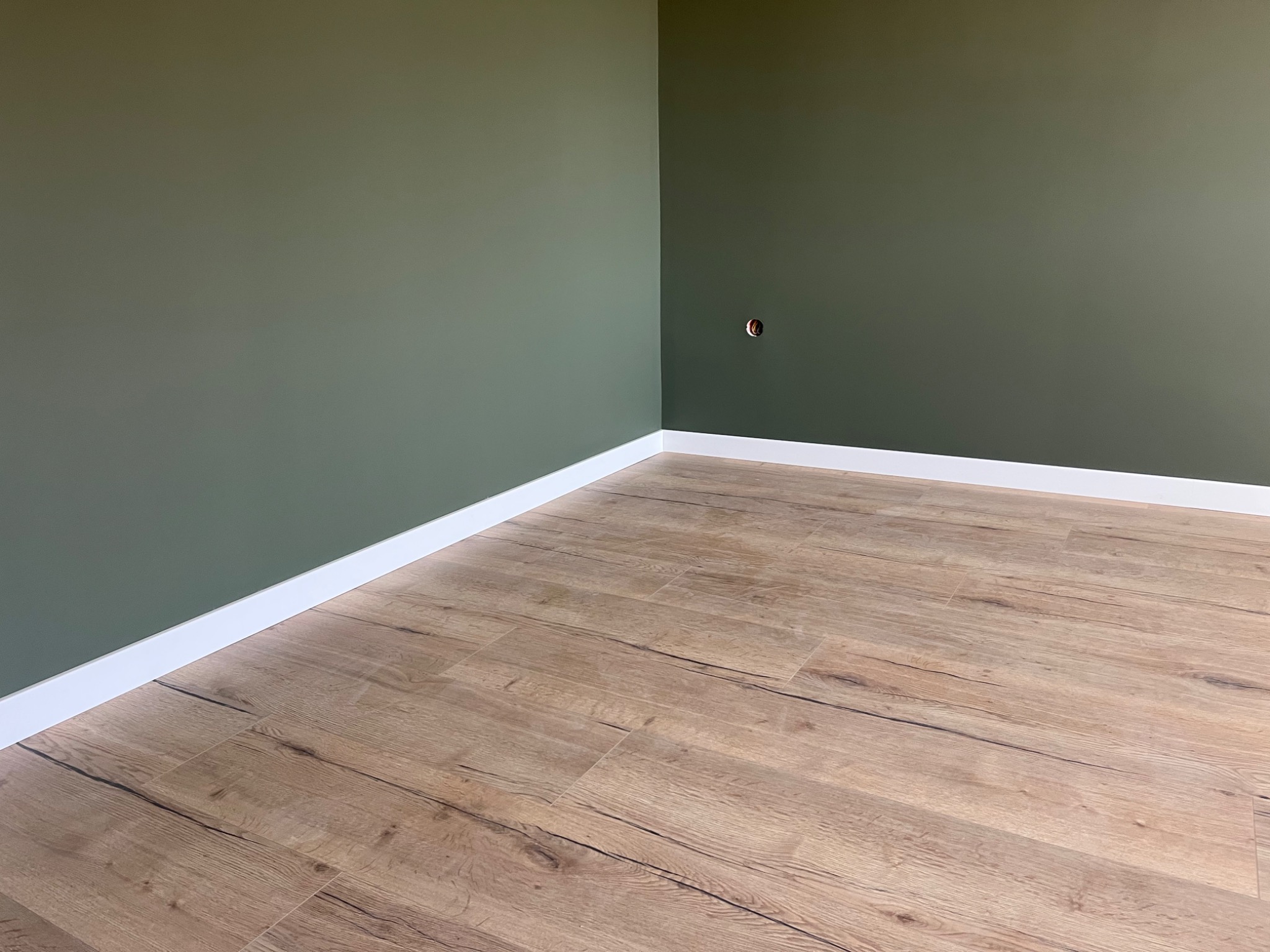 Ułożone panele podłogowe imitujące drewno, widoczne łączenia, białe listwy przypodłogowe i fragment ściany pomalowanej na oliwkowy kolor z wystającym gniazdkiem elektrycznym.