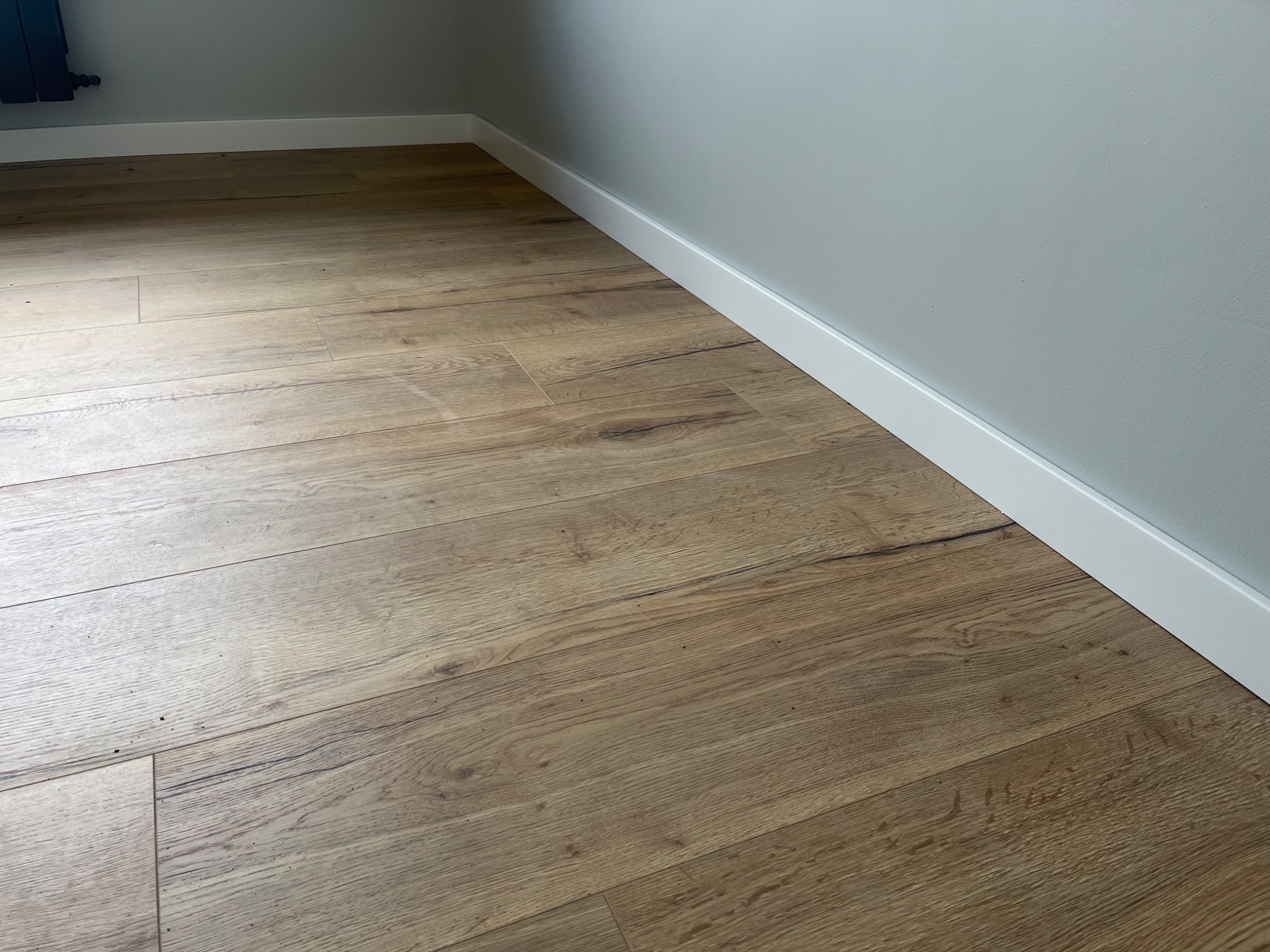 Ułożone panele podłogowe imitujące drewno, z widoczną białą listwą przypodłogową przy szarej ścianie.
