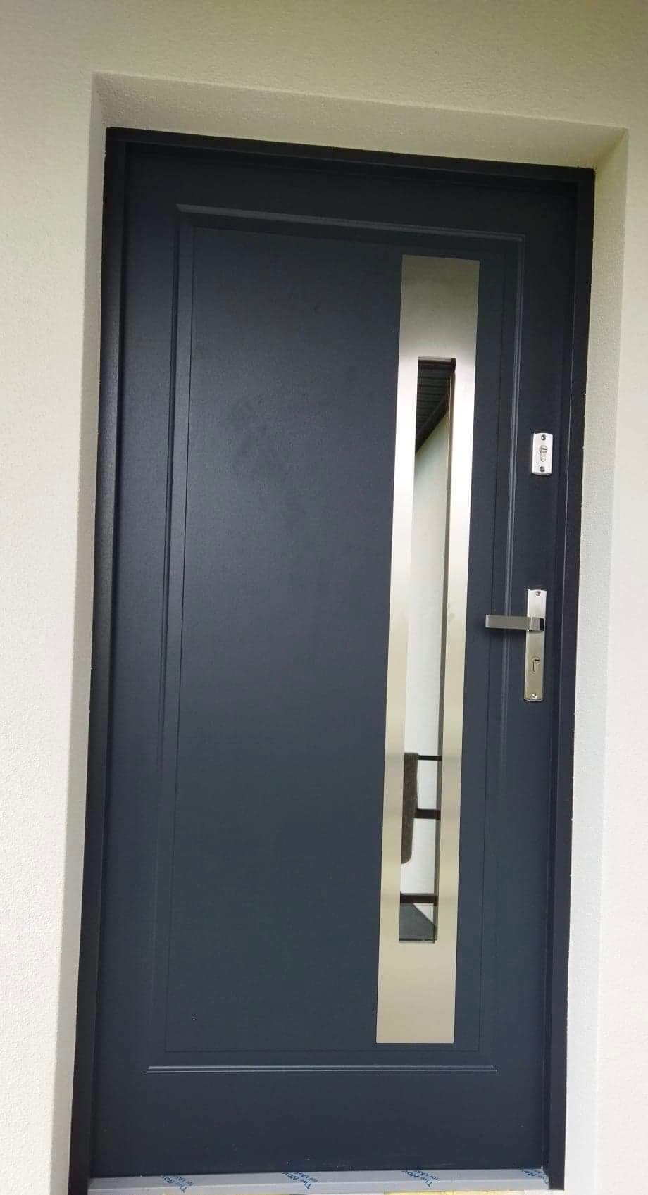 Grafitowe drzwi wejściowe z pionowym, metalowym panelem z przeszkleniem i srebrną klamką na tle białej ściany.