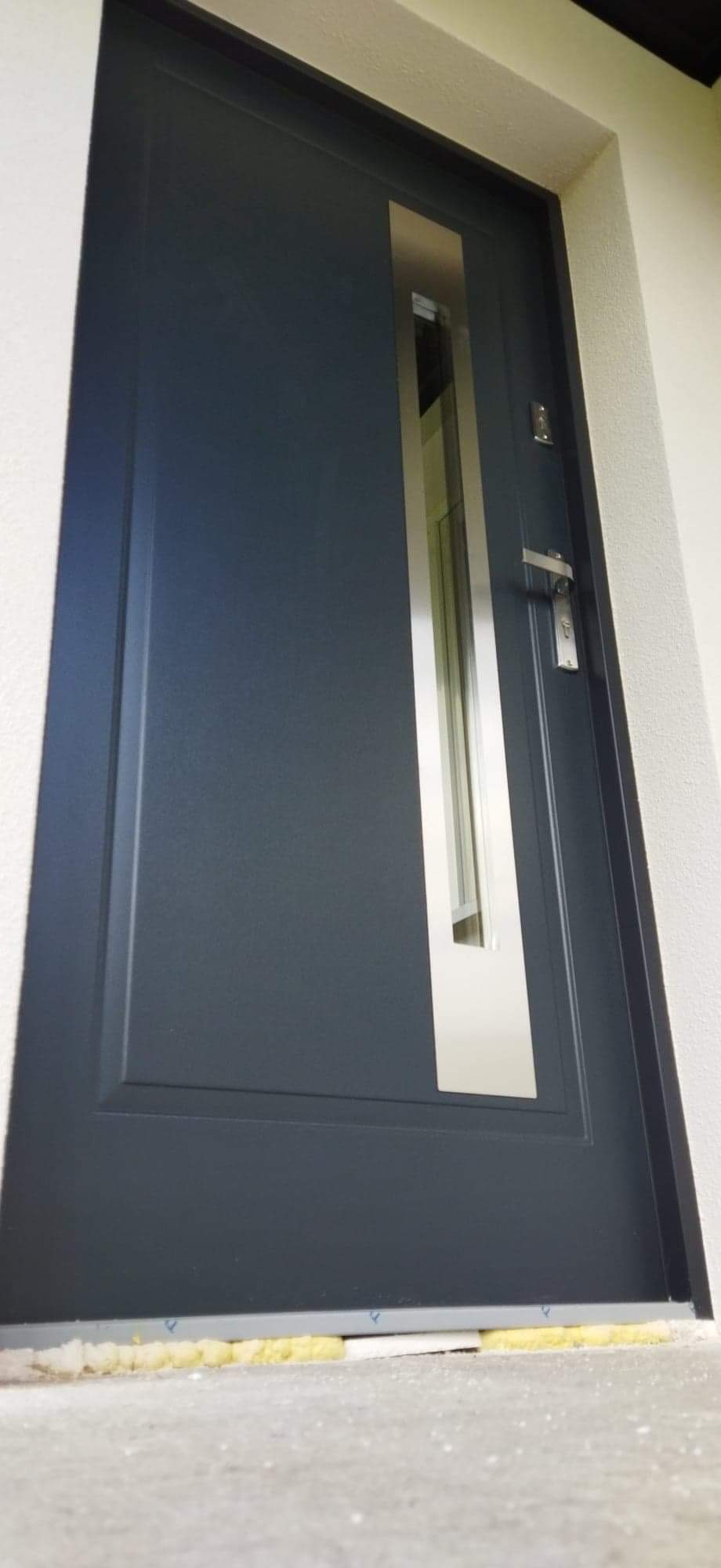 Grafitowe drzwi zewnętrzne z pionowym, wąskim przeszkleniem w stalowej ramie, widoczne ślady montażu u dołu.