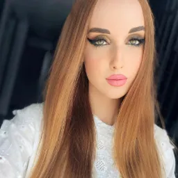 Portret kobiety z długimi, prostymi, rudymi włosami, mocnym makijażem oczu z czarną kreską i różowymi ustami, ubrana w białą bluzkę z haftem.