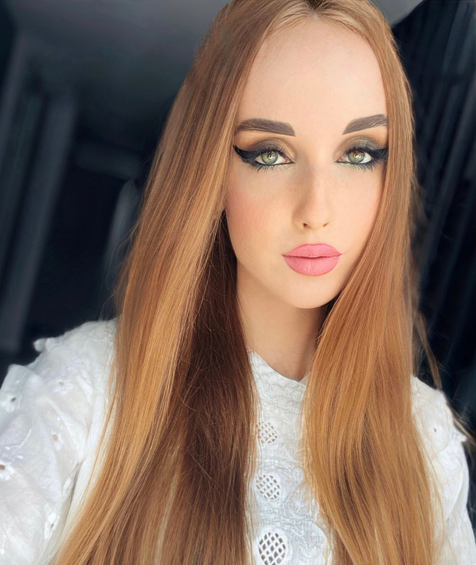 Portret kobiety z długimi, prostymi, rudymi włosami, mocnym makijażem oczu z czarną kreską i różowymi ustami, ubrana w białą bluzkę z haftem.