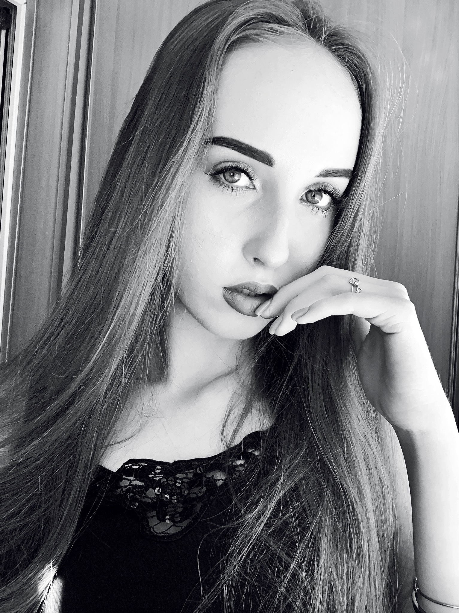 Portret kobiety w czarno-białej tonacji, z długimi włosami, delikatnym makijażem i pierścionkiem na palcu, pozującej z dłonią przy ustach.