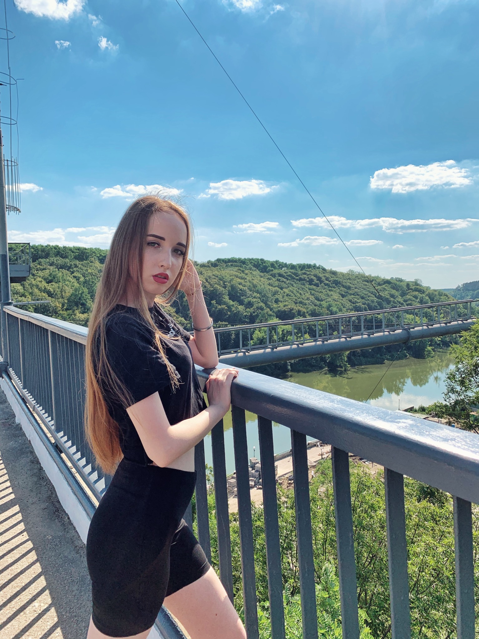 Portret młodej kobiety w czarnym stroju, opierającej się o metalową barierkę mostu linowego z widokiem na zalesione wzgórza i rzekę w słoneczny dzień.