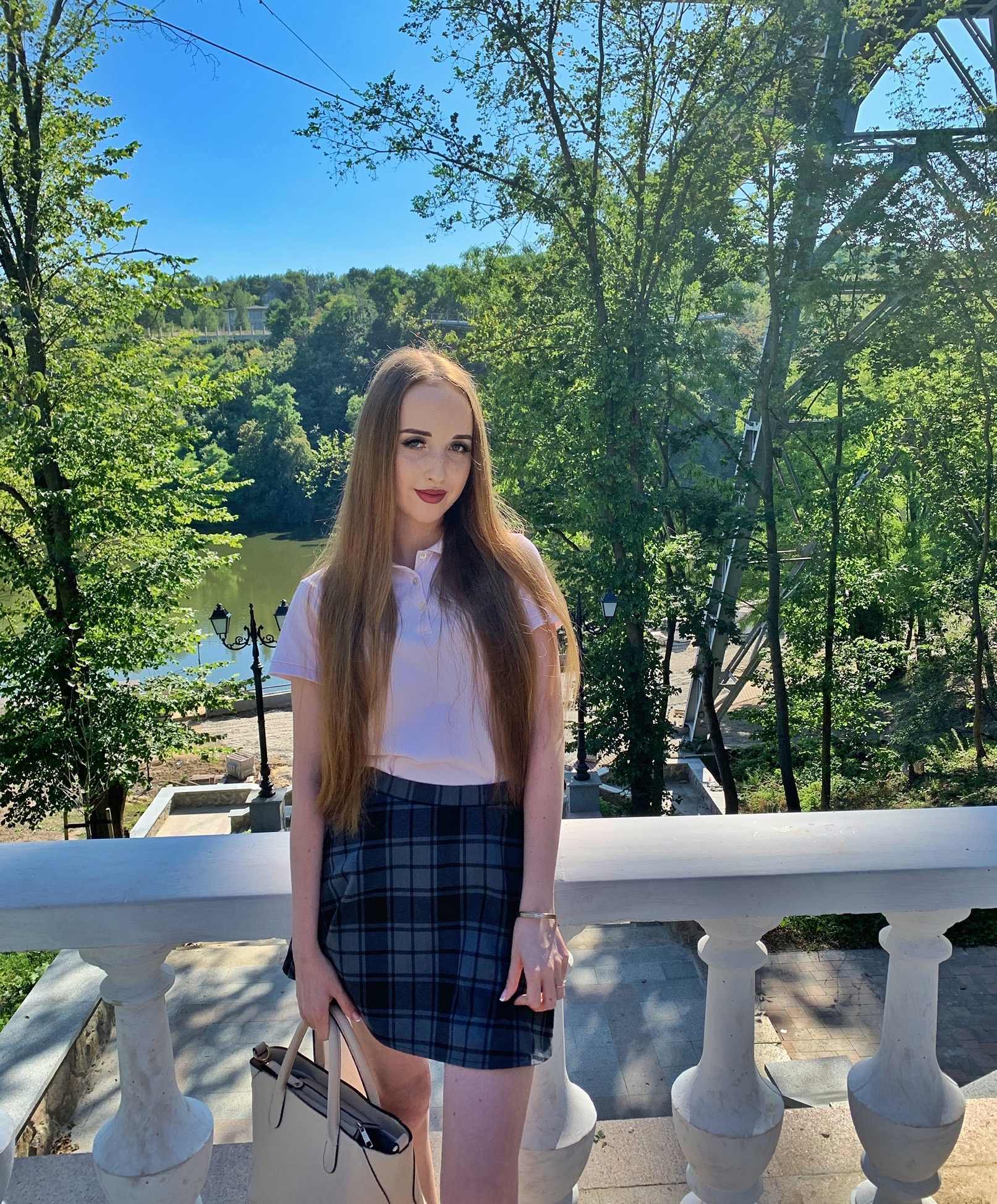 Portret kobiety na tle parku z jeziorem, w słoneczny dzień. Ubrana w spódnicę w kratę i bluzkę polo, trzyma beżową torbę. W tle widoczny most i zieleń.