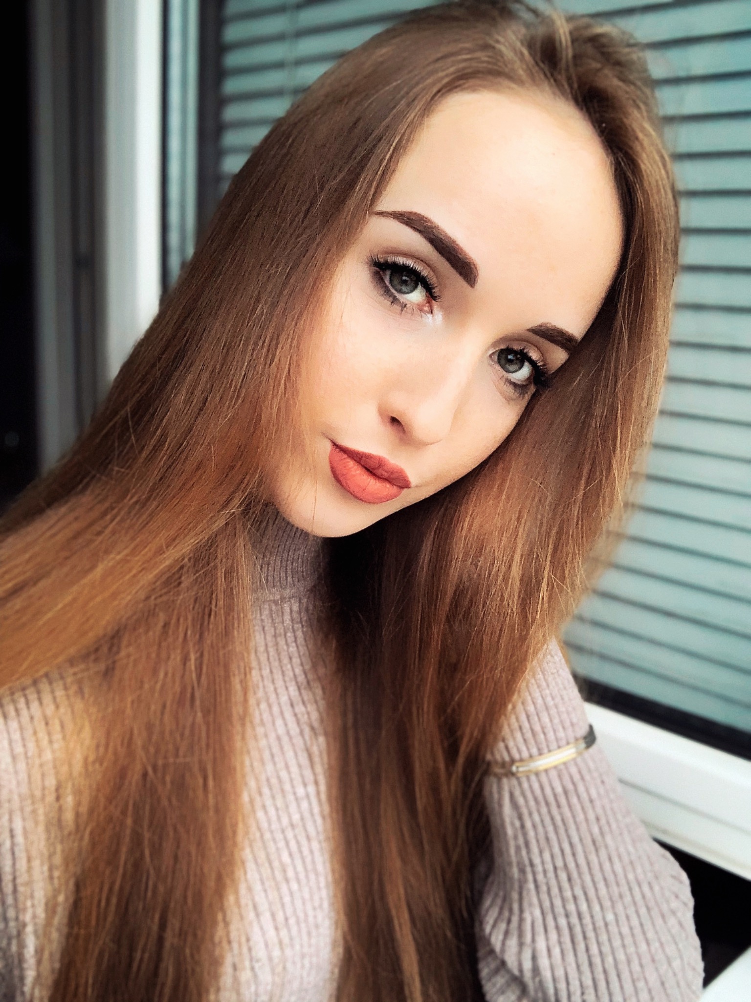 Portret młodej kobiety z długimi, rudymi włosami, delikatnym makijażem i bransoletką na ręce, pozującej przy oknie z żaluzjami.