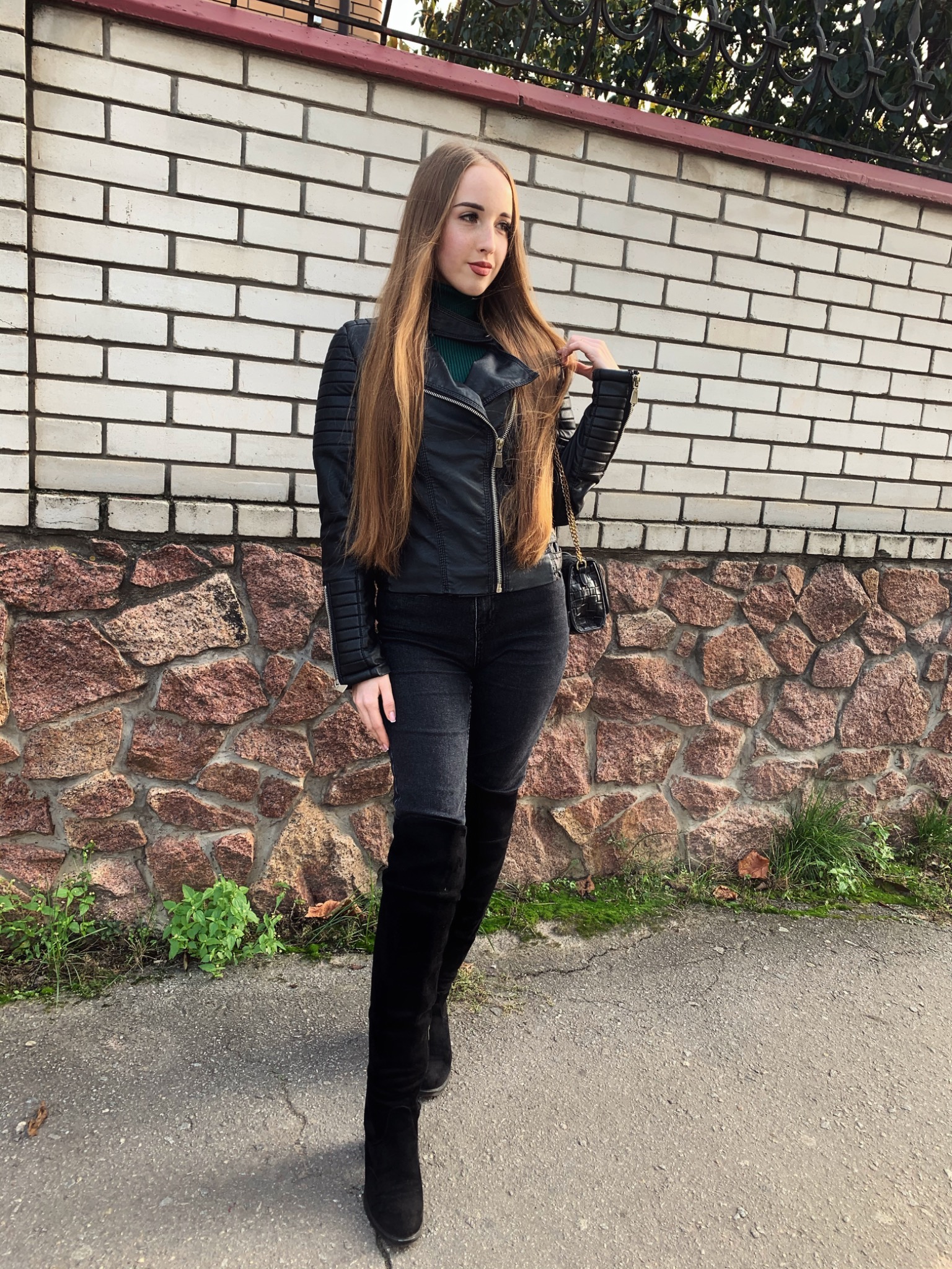 Kobieta w czarnej skórzanej kurtce, dżinsach i kozakach za kolano pozuje na tle ściany z cegieł i kamienia.
