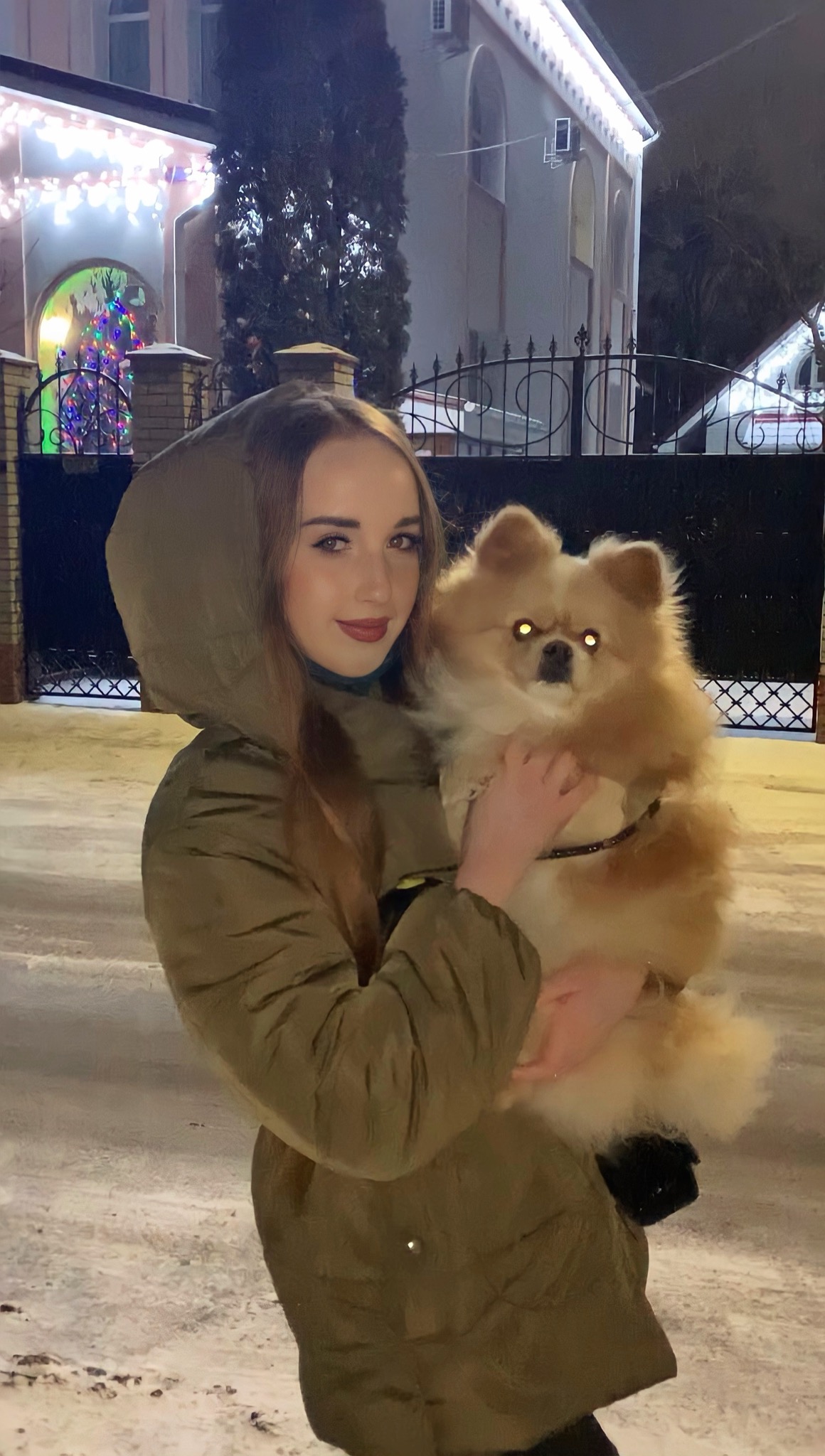 Uśmiechnięta kobieta w zimowej kurtce z kapturem trzyma na rękach puszystego szpica miniaturowego na tle ośnieżonej ulicy i domu z dekoracjami świątecznymi.