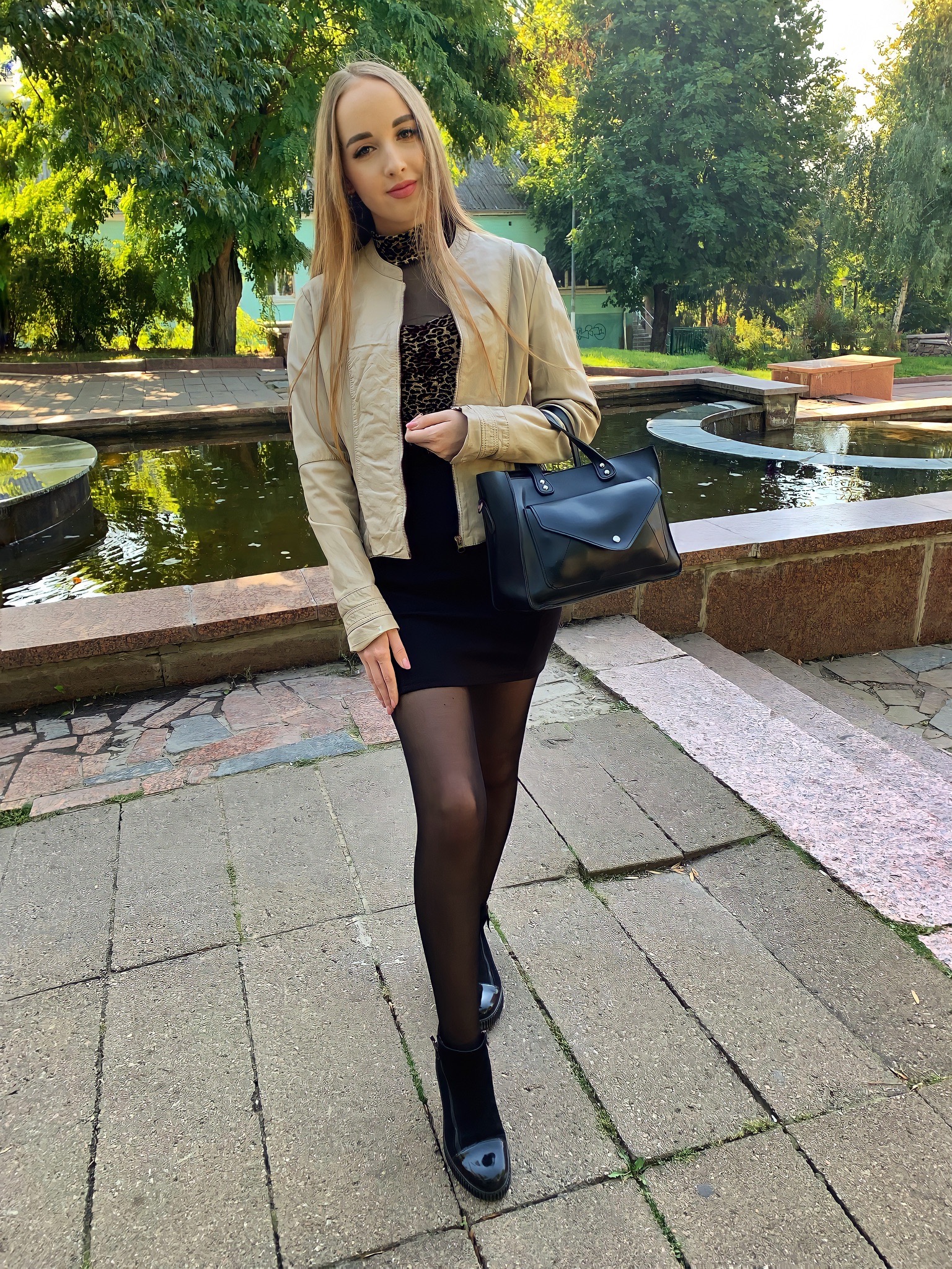 Kobieta w czarnej sukience i beżowej kurtce pozuje na tle fontanny w parku. W ręku trzyma czarną torebkę. Ujęcie z dołu, widoczne drzewa i budynki w tle.