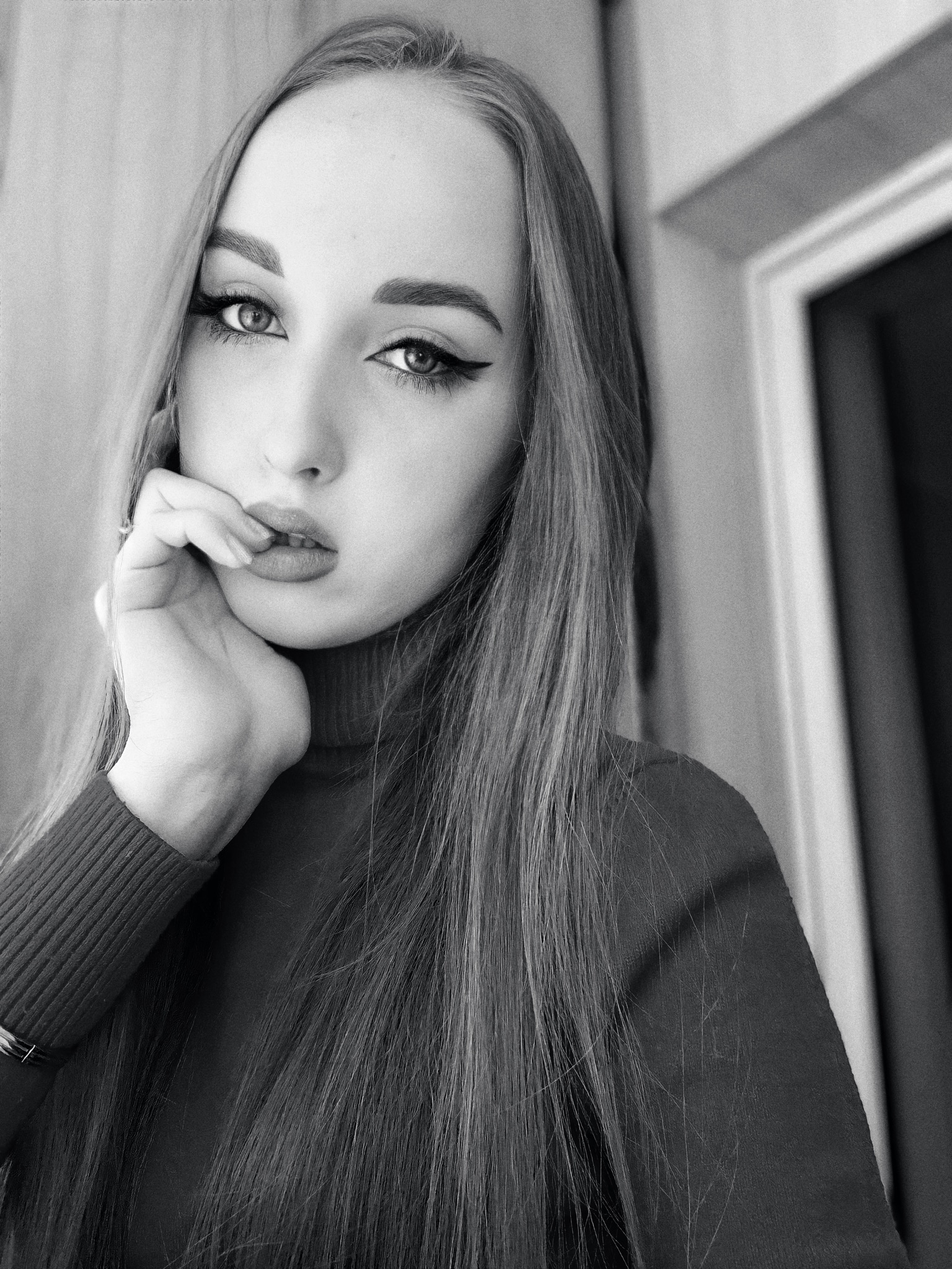 Portret kobiety z długimi włosami, mocnym makijażem oczu i golfem, w czerni i bieli, z ręką przy ustach.