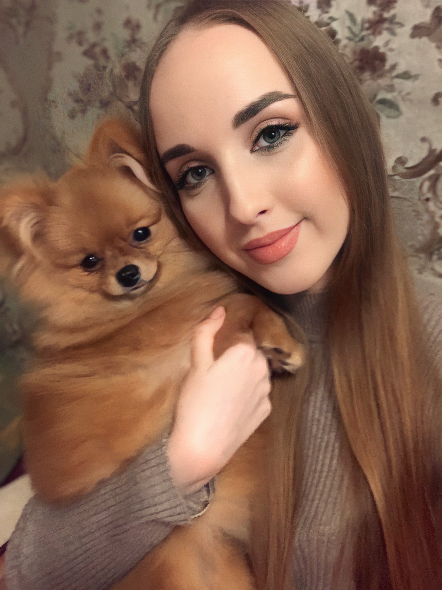 Portret młodej kobiety z długimi, prostymi włosami w odcieniach blondu, trzymającej na rękach małego, rudego szpica miniaturowego na tle ściany z kwiecistym wzorem