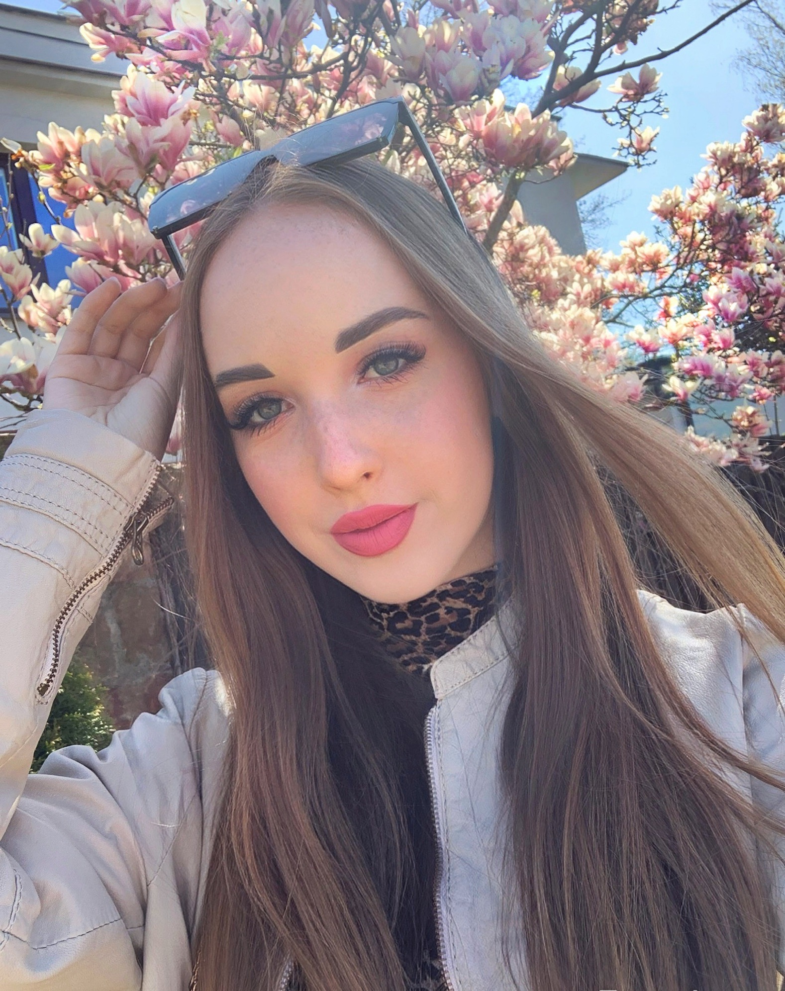 Portret młodej kobiety z okularami na głowie, na tle kwitnącej magnolii w słoneczny dzień. Uśmiech, jasna skórzana kurtka, wzorzysta bluzka, naturalny makijaż.