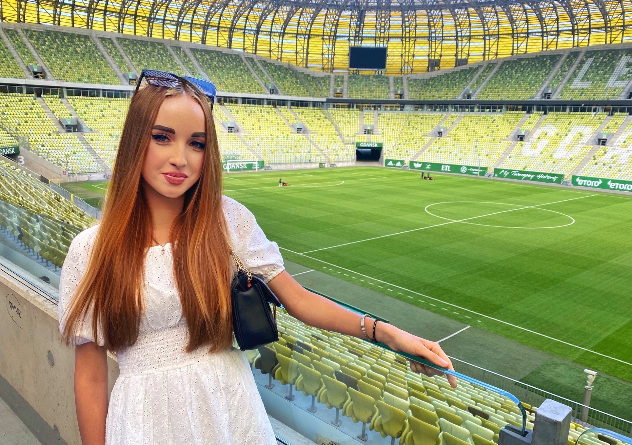 Młoda kobieta w białej sukience pozuje na tle zielonej murawy stadionu w Gdańsku, z widocznymi trybunami i konstrukcją dachu.