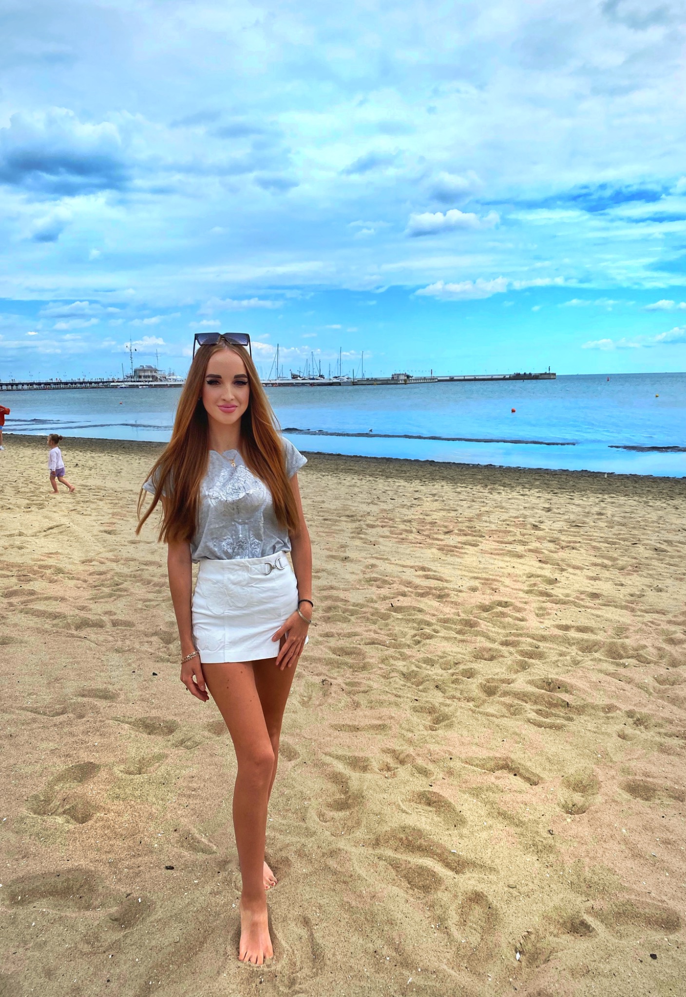 Młoda kobieta w białej spódnicy i szarej bluzce pozuje na piaszczystej plaży z widokiem na molo i morze w tle.