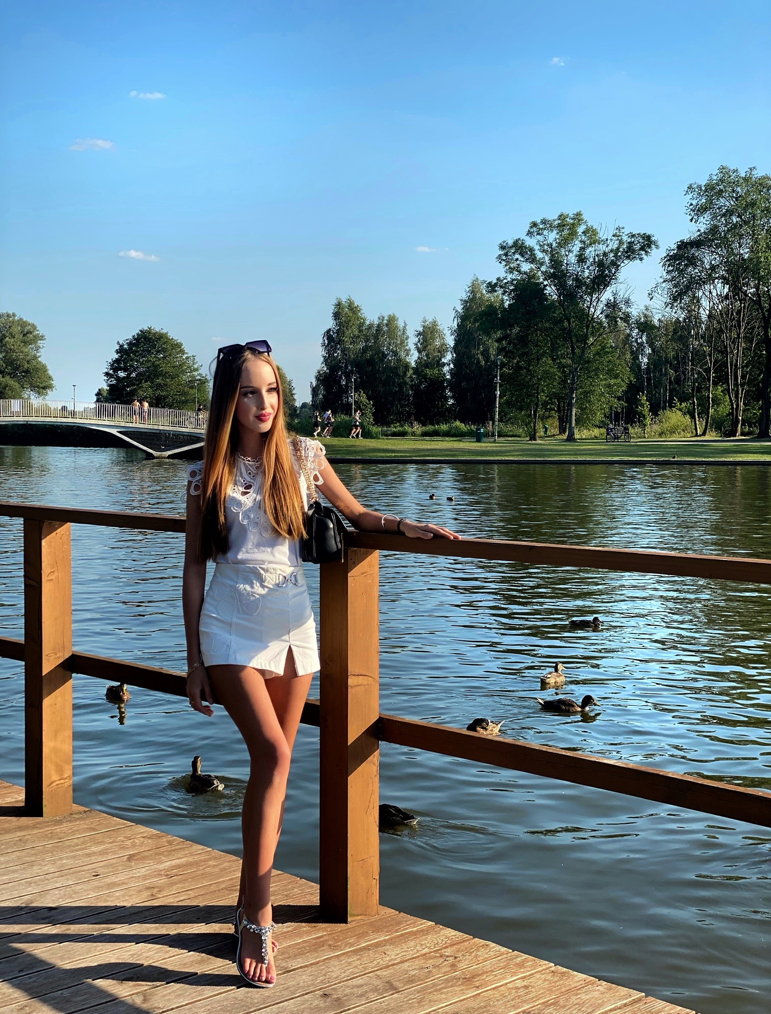 Kobieta w białym stroju pozuje na drewnianym pomoście nad jeziorem z kaczkami, w tle widoczny park i most.