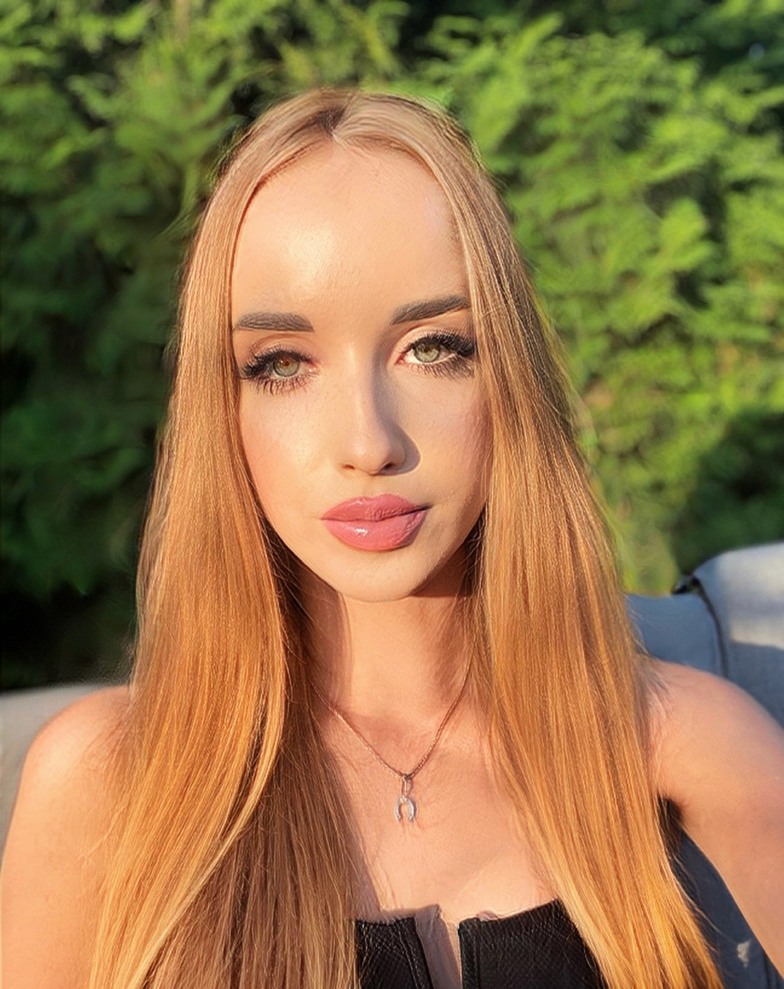 Portret młodej kobiety z długimi, prostymi, rudymi włosami, delikatnym makijażem i zawieszką w kształcie podkowy na szyi, na tle zieleni.