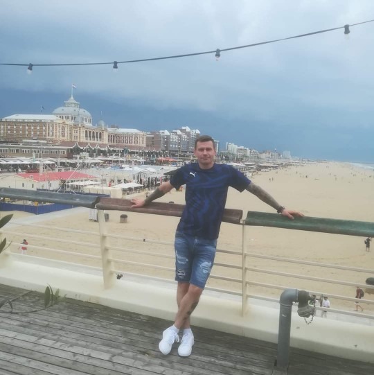 Mężczyzna oparty o barierkę drewnianego pomostu z widokiem na plażę i zabudowę miejską w oddali, pochmurne niebo.