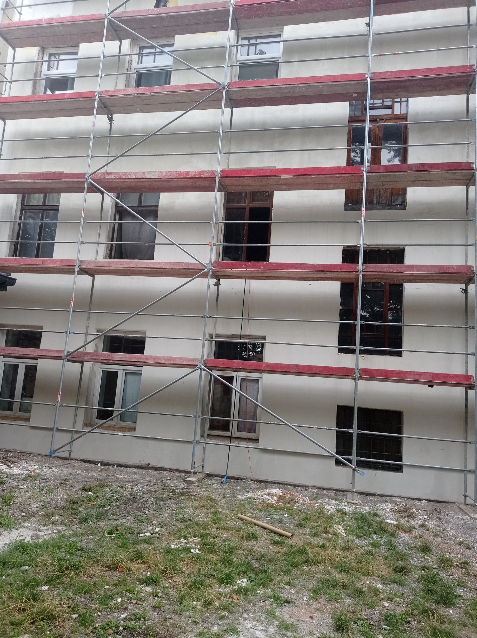 Budynek wielorodzinny w trakcie prac elewacyjnych, z widocznym rusztowaniem i świeżo nałożonym tynkiem w kolorze złamanej bieli, stare okna z brązowymi ramami.