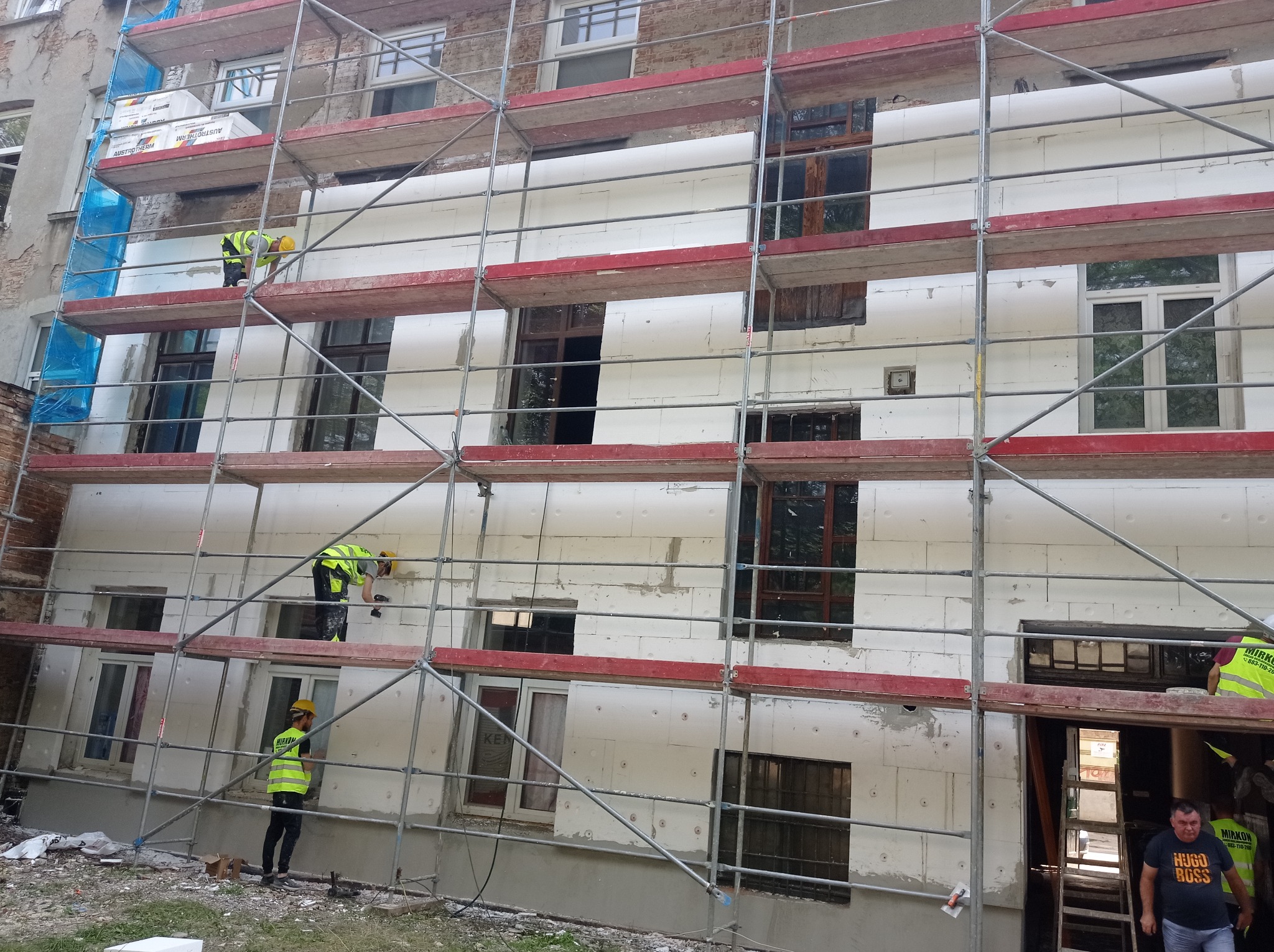 Pracownicy w żółtych kamizelkach montują białe płyty izolacyjne na elewacji budynku z cegły, widoczne rusztowania.