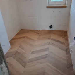 Ułożona w jodełkę podłoga z płytek imitujących drewno w jasnej łazience z białymi kafelkami na ścianach, widoczne wystające rury i okno.