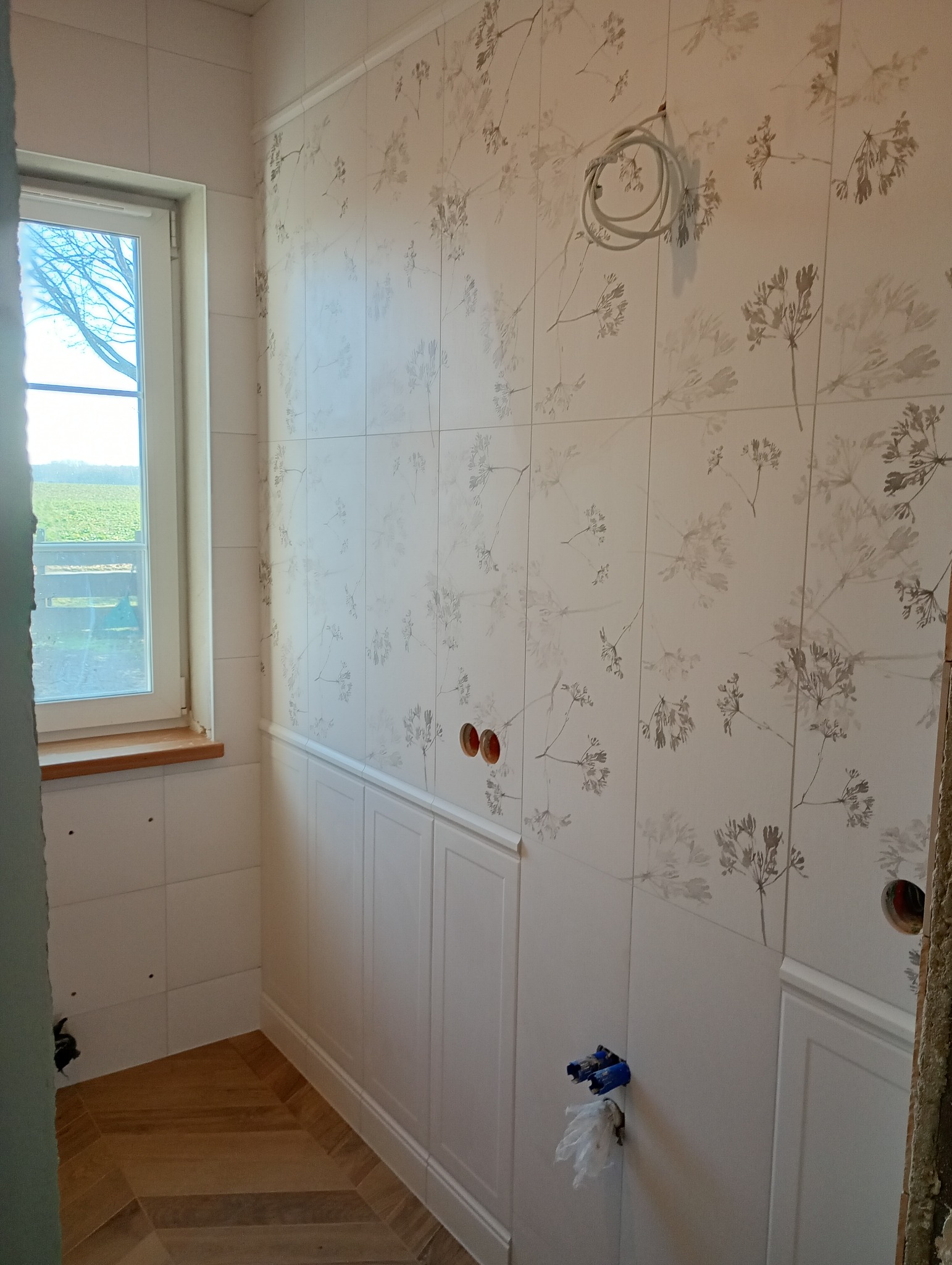 Wnętrze łazienki w trakcie prac: ułożone płytki z motywem roślinnym na ścianach, dolna część ściany wykończona białą boazerią, widoczne wystające kable i rury, podłoga wyłożona parkietem w jodełkę,...