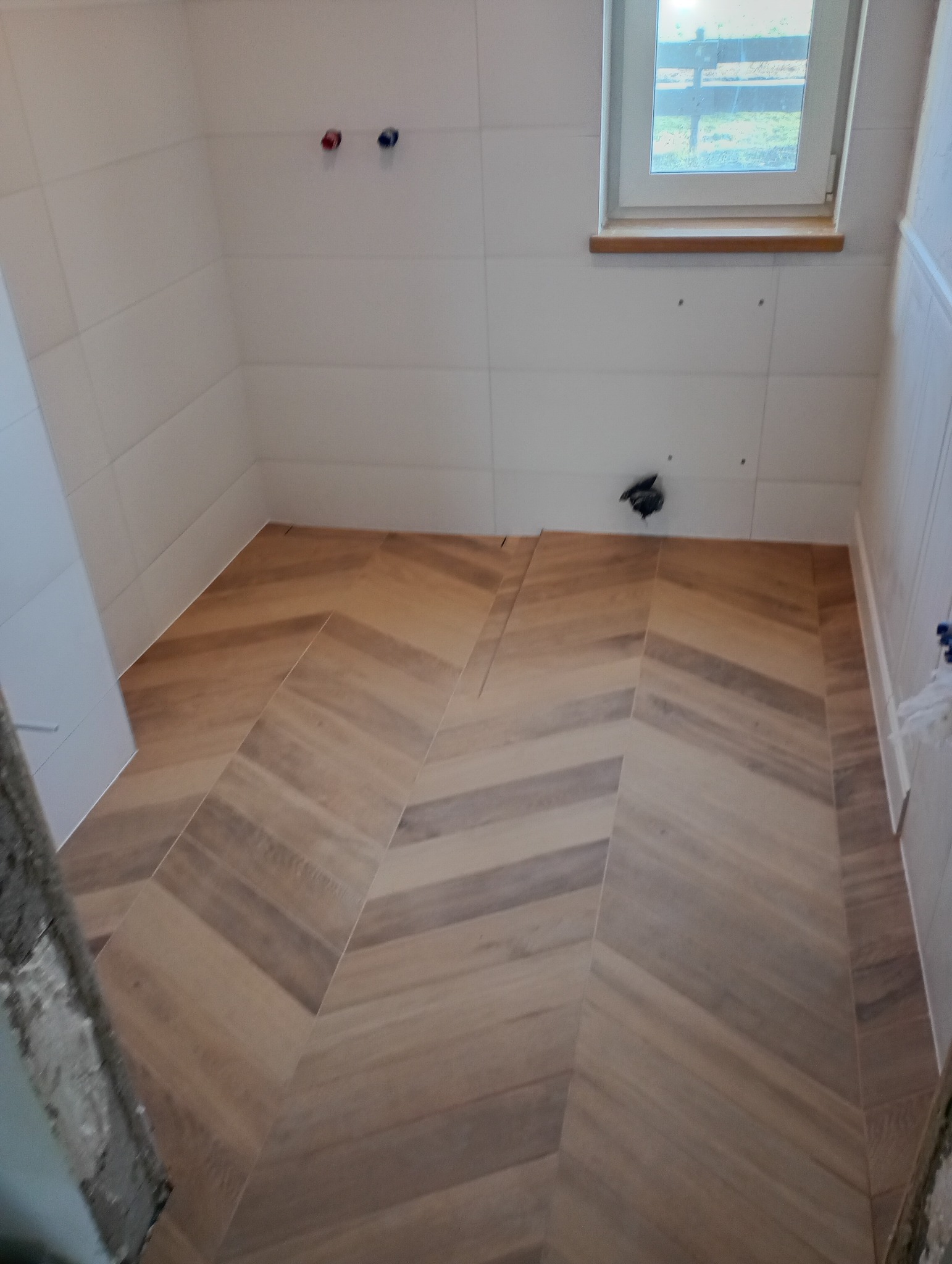 Ułożona w jodełkę podłoga z płytek imitujących drewno w jasnej łazience z białymi kafelkami na ścianach, widoczne wystające rury i okno.