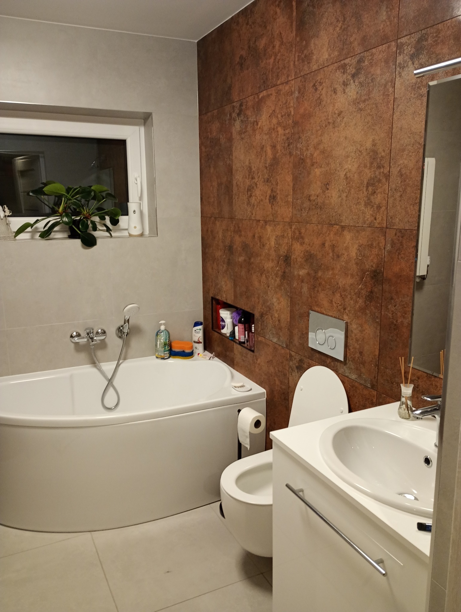 Nowoczesna łazienka z wanną narożną, umywalką nablatową i toaletą podwieszaną, wykończona płytkami w odcieniach brązu i beżu, z rośliną doniczkową na parapecie okna.