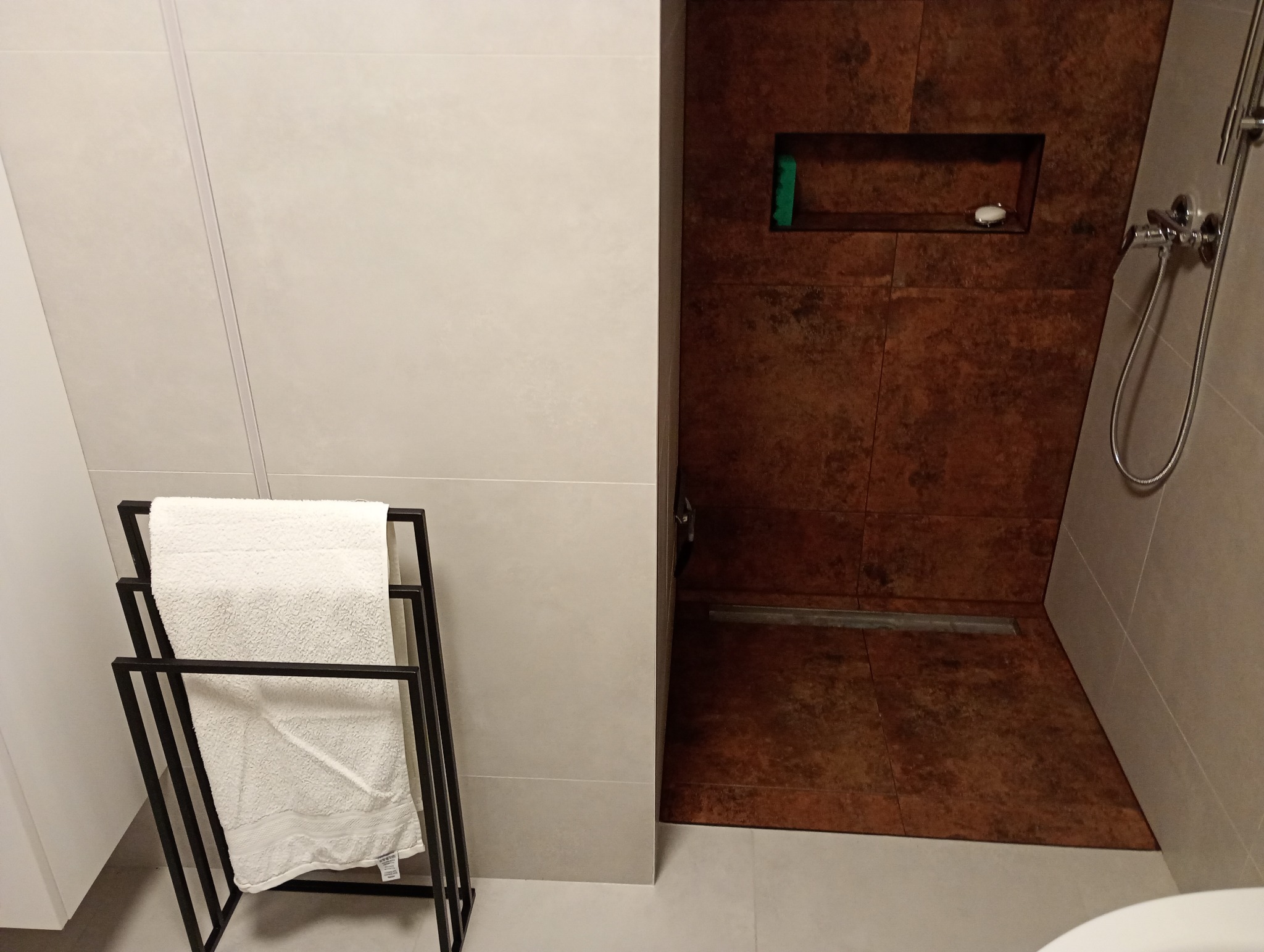 Nowoczesna łazienka z brązowymi płytkami imitującymi rdzę w kabinie prysznicowej typu walk-in i jasnymi płytkami na pozostałych ścianach, widoczny odpływ liniowy i minimalistyczny wieszak...