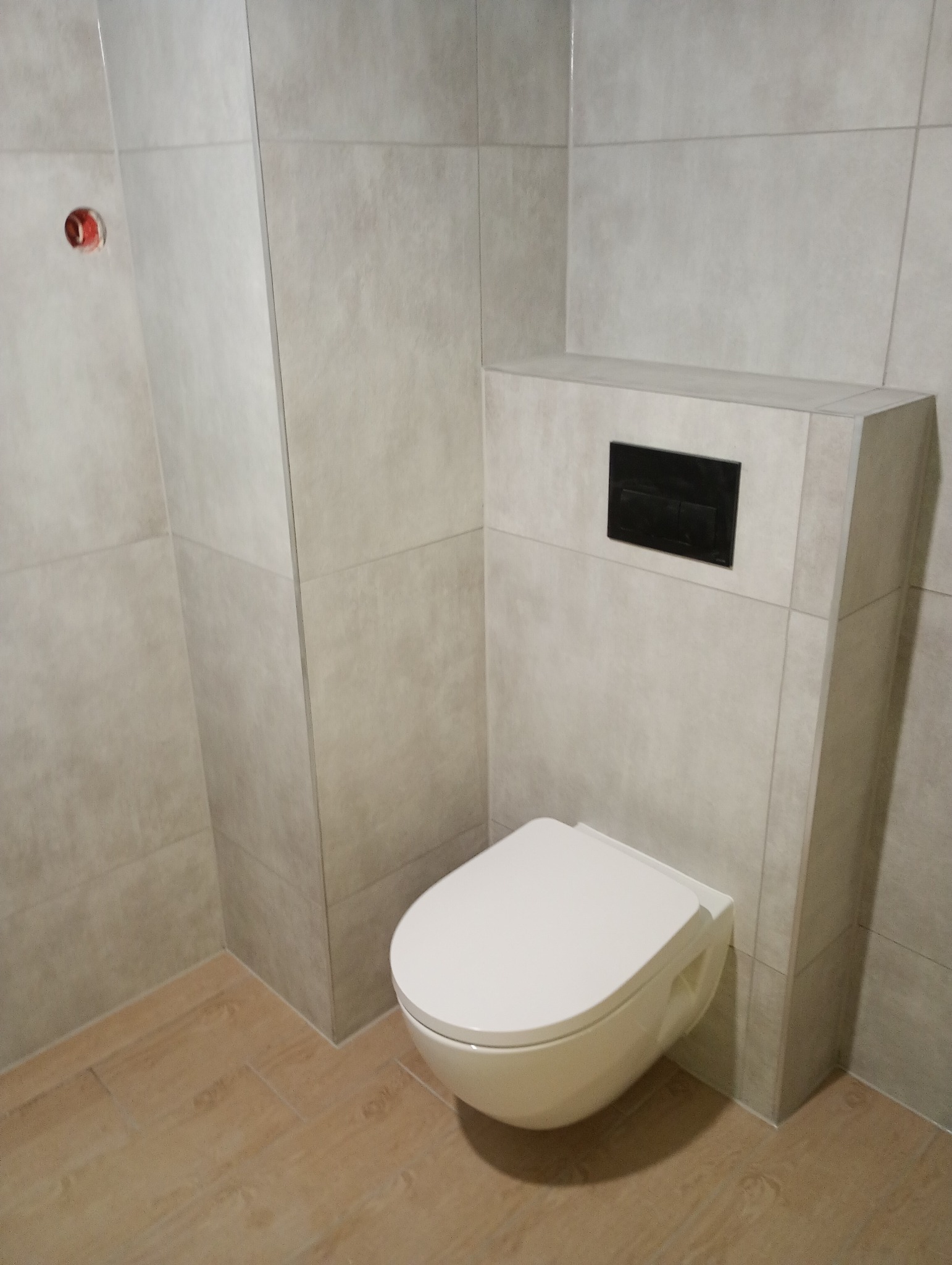 Nowoczesna, podwieszana toaleta z czarnym przyciskiem spłukującym, obudowana płytkami w odcieniach szarości, z podłogą imitującą drewno, widoczny fragment instalacji elektrycznej w ścianie.