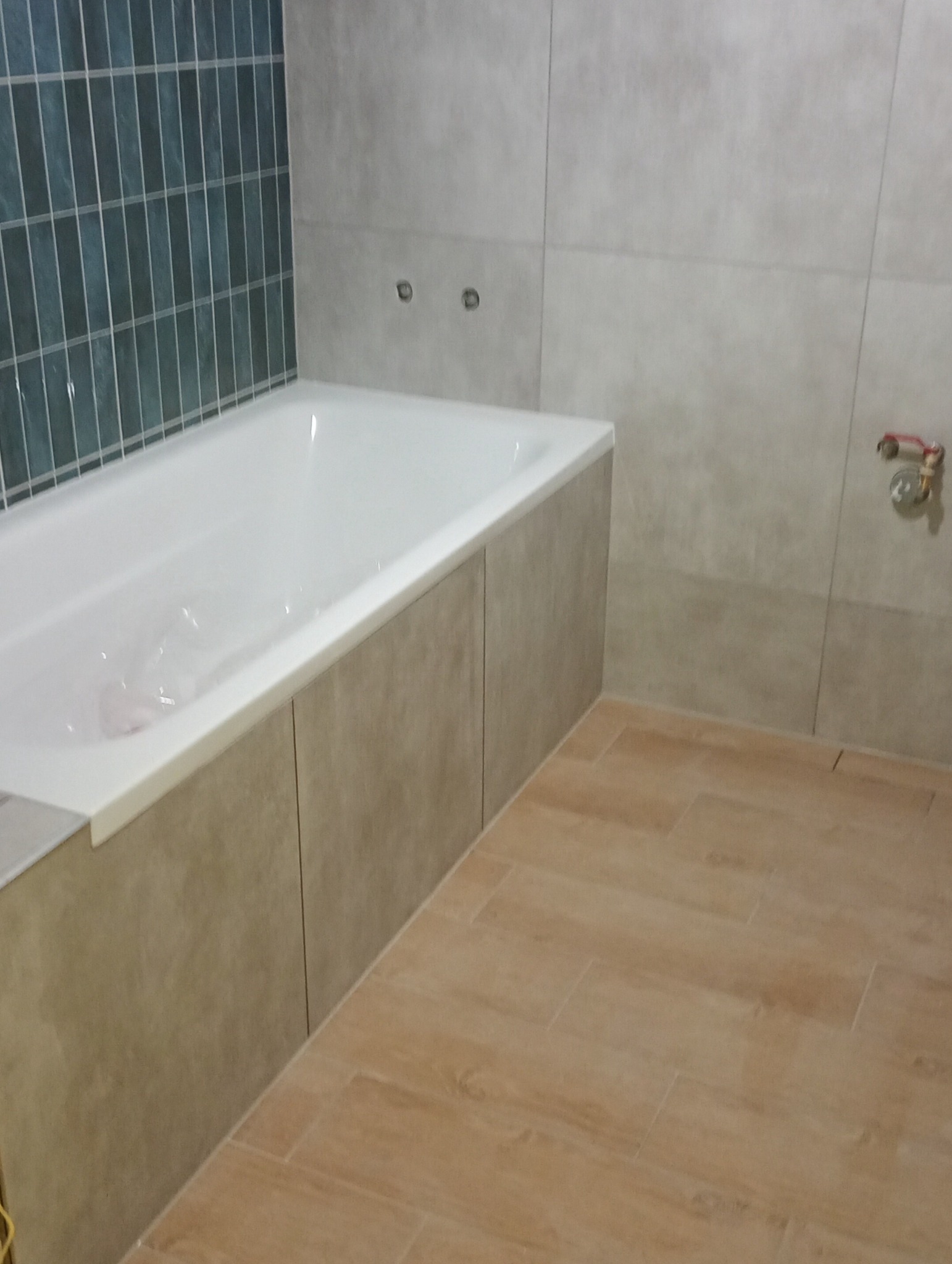 Wanna zabudowana panelami w odcieniach beżu, z boku ściana wyłożona płytkami w kolorze szarym, a na ścianie naprzeciwko wanny pionowe pasy płytek w odcieniach zieleni i szarości, podłoga wyłożona...