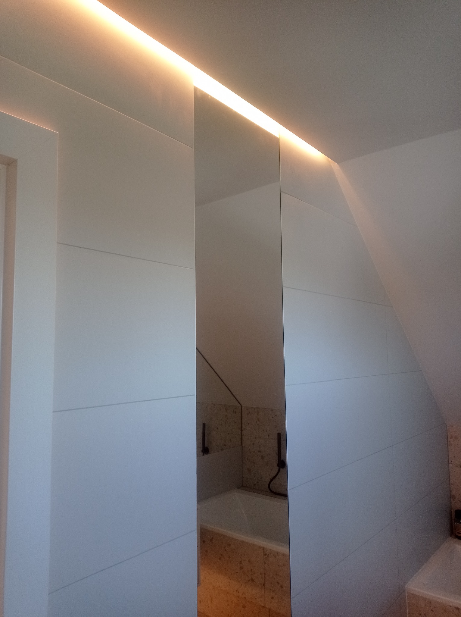 Nowoczesna łazienka z białymi płytkami i ukrytym oświetleniem LED, widoczna w lustrzanym odbiciu wanna z czarną armaturą i beżowymi płytkami.