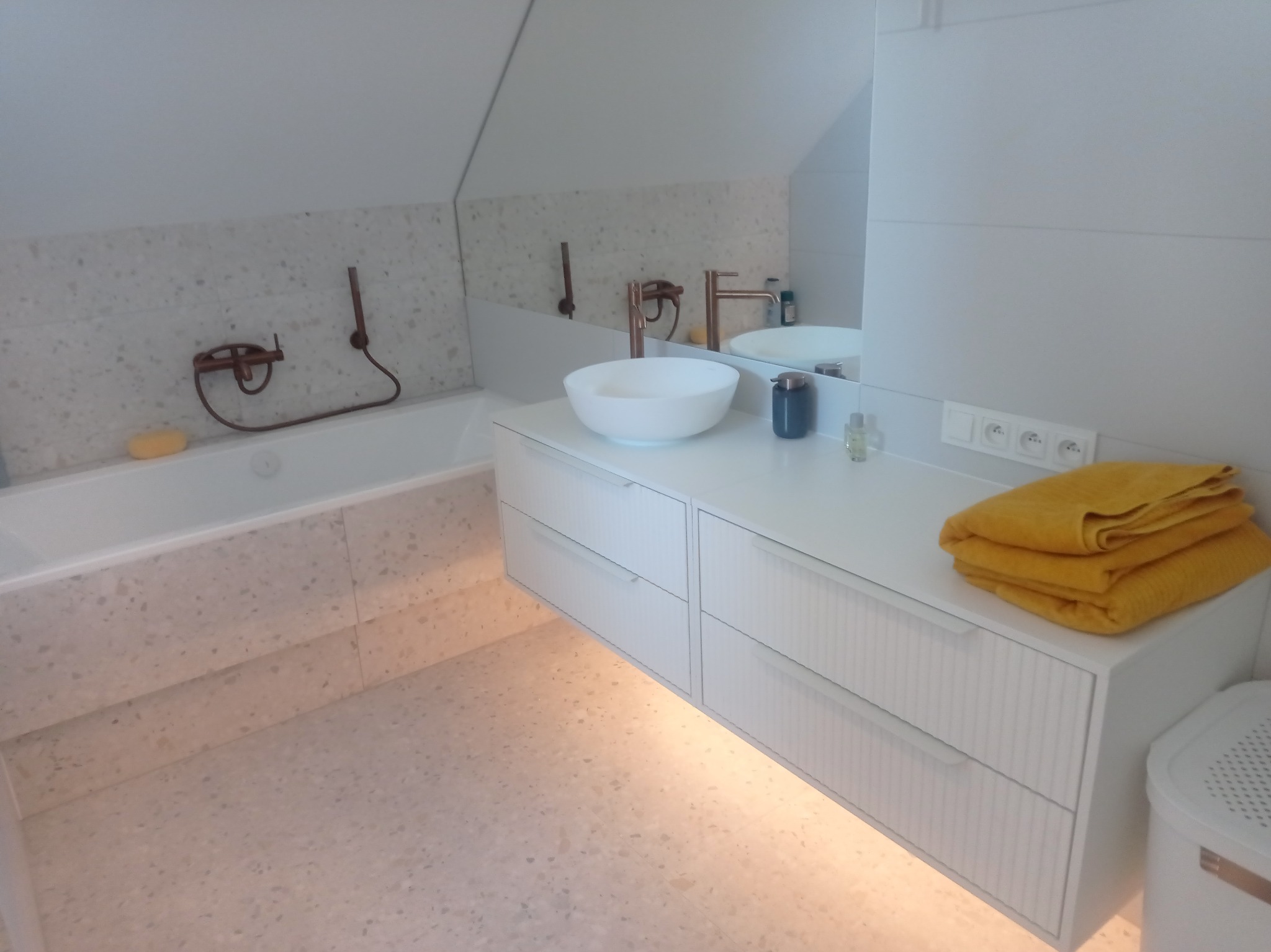 Nowoczesna łazienka na poddaszu z wanną, umywalką nablatową, szafką z szufladami z ryflowanymi frontami, jasną mozaikową posadzką i ścianami oraz miedzianą armaturą.