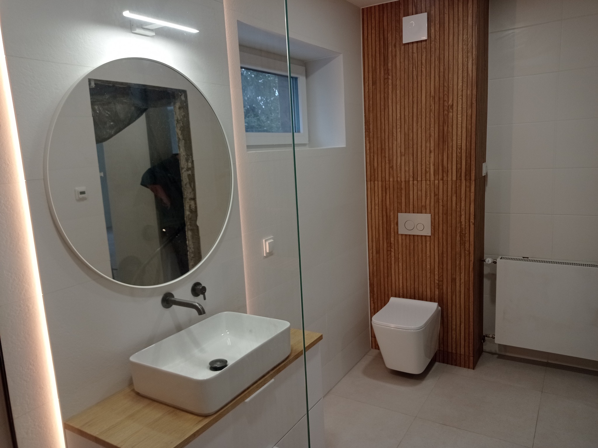 Nowoczesna łazienka z okrągłym lustrem z podświetleniem LED, prostokątną umywalką nablatową, drewnianym akcentem pod umywalką i na ścianie za toaletą, oraz grzejnikiem.