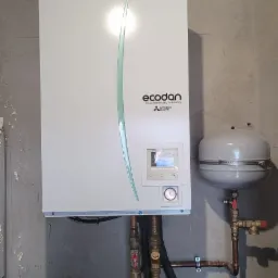 Biała jednostka Ecodan Mitsubishi Electric zamontowana na szarej, surowej ścianie z widocznymi rurami miedzianymi i czarnymi izolowanymi, obok szary zbiornik wyrównawczy i instalacja elektryczna...