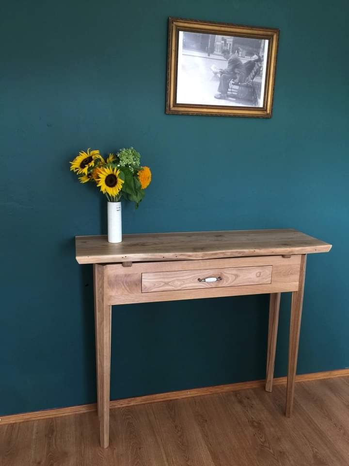 Drewniana konsola z szufladą i ceramicznym uchwytem, na tle ciemnoturkusowej ściany z obrazem w złotej ramie i wazonem ze słonecznikami.
