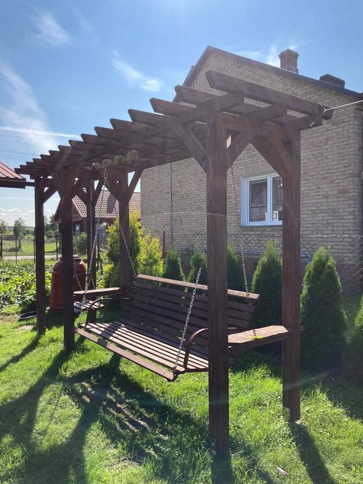 Drewniana huśtawka ogrodowa pod zadaszeniem z belek, zawieszona na łańcuchach, na tle zielonego trawnika i domu z cegły