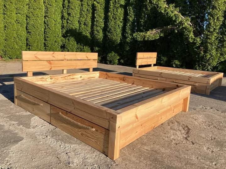 Dwa drewniane łóżka z szufladami i zagłówkami, wykonane z jasnego drewna, ustawione na zewnątrz na betonowej powierzchni, z zielonymi krzewami w tle.