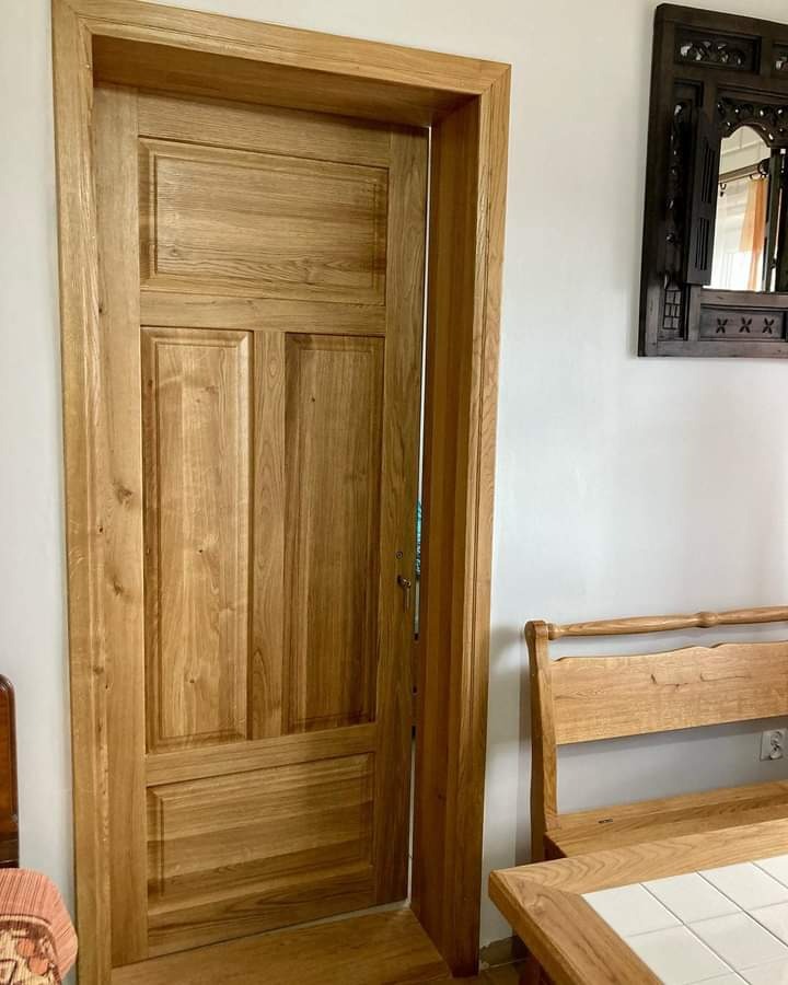 Otwarta, drewniana, wewnętrzna  drzwi z ościeżnicą, widoczny fragment mebla z jasnego drewna i dekoracyjne lustro na białej ścianie.