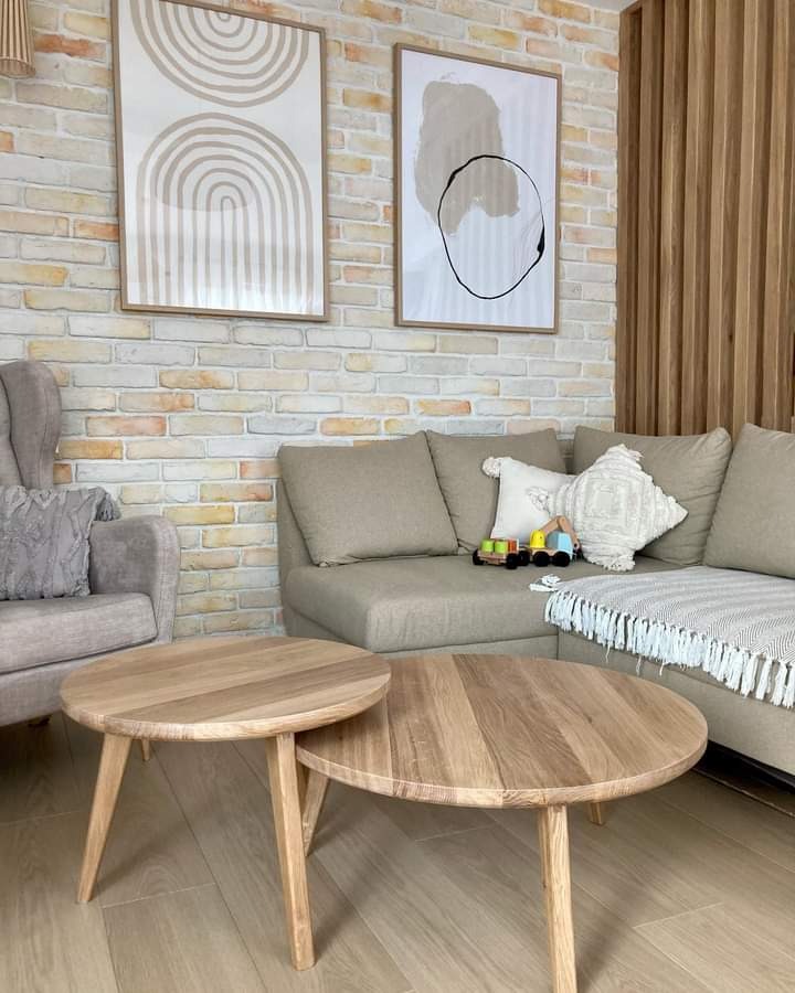 Dwa okrągłe stoliki kawowe z jasnego drewna na tle ceglanej ściany z dekoracyjnymi obrazami, szarej sofy i fotela