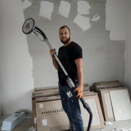 M.M-complexRem - Mężczyzna z uśmiechem trzyma szlifierkę do gładzi na tle częściowo wyszlifowanej ściany, obok stos kartonów płytek ceramicznych.