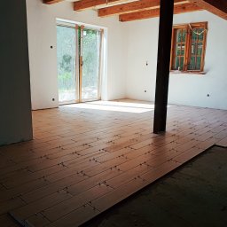 M.M-complexRem - Układanie płytek podłogowych imitujących drewno w jasnym pomieszczeniu z drewnianym stropem i oknami, widoczne krzyżyki dystansowe i częściowo ułożona podłoga.