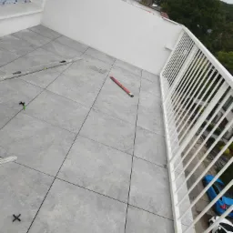 Szare płytki gresowe ułożone na balkonie z białymi barierkami, widoczne poziomice, krzyżyki dystansowe, w tle zabudowa miejska i zieleń