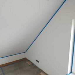 M.M-complexRem - Poddasze przygotowane do malowania: ściany zabezpieczone niebieską taśmą malarską, podłoga przykryta folią ochronną, widoczne gniazdka elektryczne i fragment drewnianej belki stropowej.