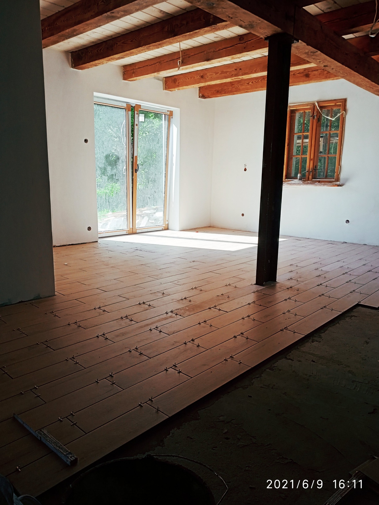 Układanie płytek podłogowych imitujących drewno w jasnym pomieszczeniu z drewnianym stropem i oknami, widoczne krzyżyki dystansowe i częściowo ułożona podłoga.