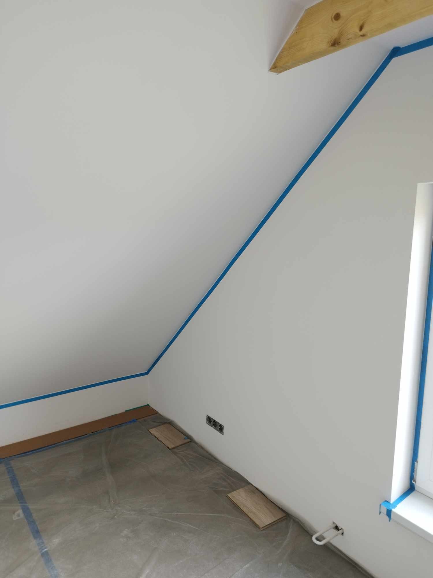 Poddasze przygotowane do malowania: ściany zabezpieczone niebieską taśmą malarską, podłoga przykryta folią ochronną, widoczne gniazdka elektryczne i fragment drewnianej belki stropowej.