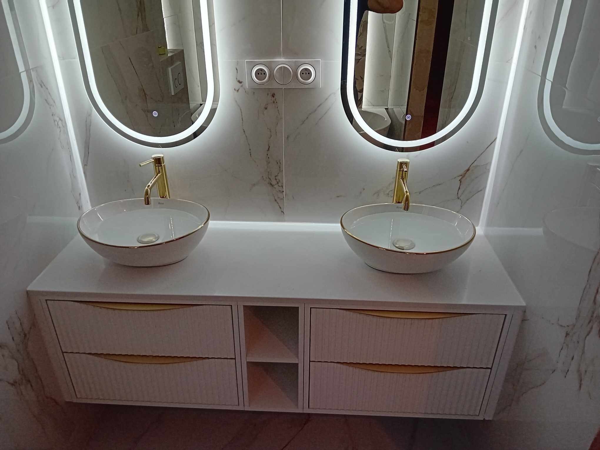 Nowoczesna łazienka z podwójną umywalką nablatową, złotymi bateriami i podświetlanymi lustrami owalnymi na marmurowej ścianie.