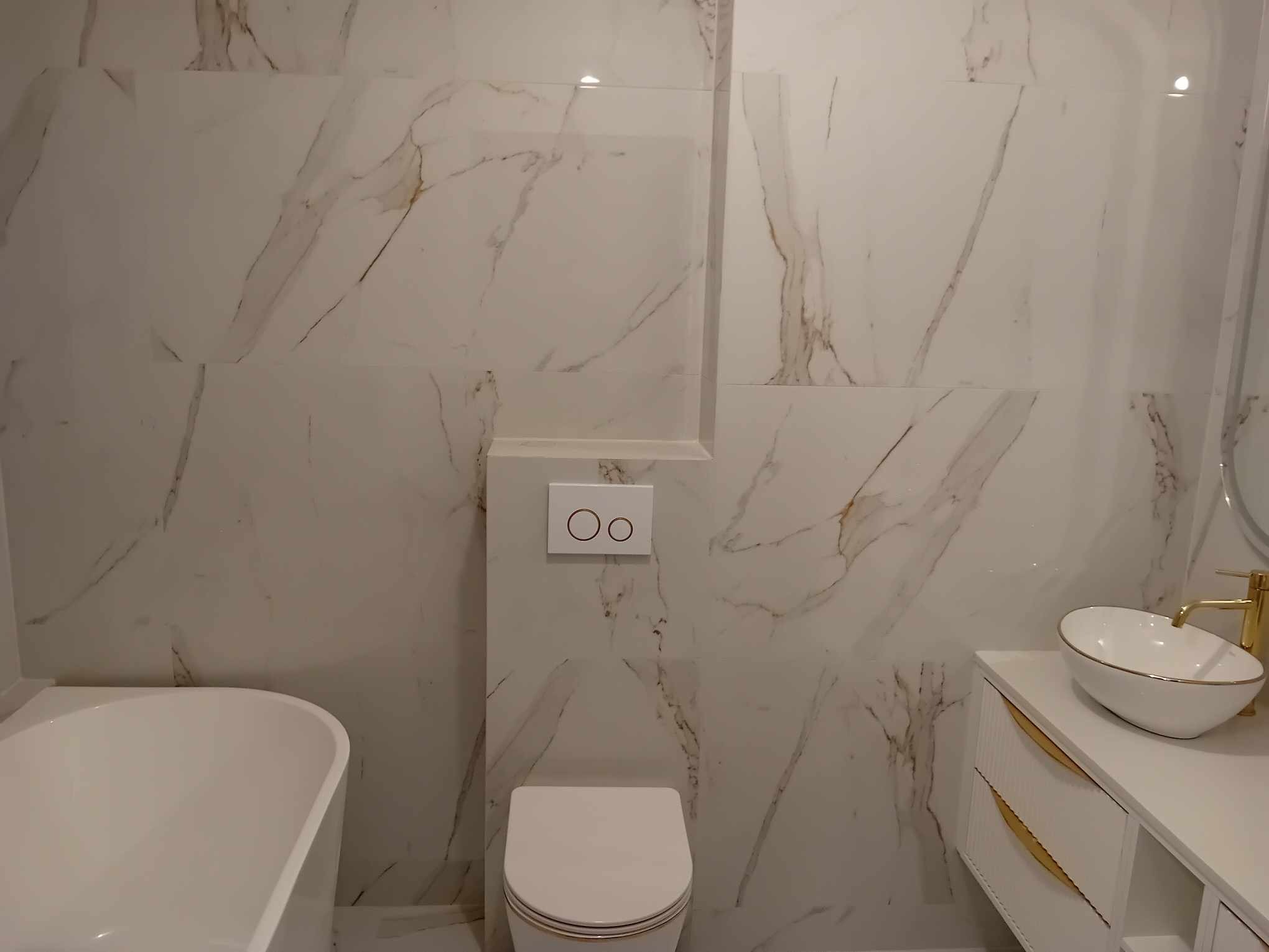 Nowoczesna łazienka z białymi płytkami imitującymi marmur, owalną umywalką nablatową ze złotą armaturą oraz podwieszaną toaletą.