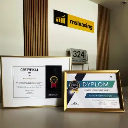 Certyfikat i dyplom w złotych ramkach na stole, logo MSLeasing na ścianie w tle, adres Bohaterów Monte Cassino 324.